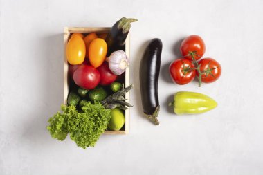 Delivery of fresh vegetables. Eggplants, beans, tomatoes, cucumbers, peppers, lettuce, garlic lie in a wooden box on a gray background, vegetables lie nearby. Flat lay, top view, knolling. clipart