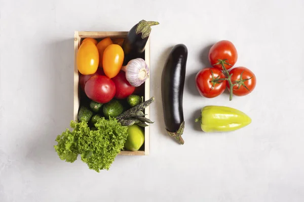 Delivery of fresh vegetables. Eggplants, beans, tomatoes, cucumbers, peppers, lettuce, garlic lie in a wooden box on a gray background, vegetables lie nearby. Flat lay, top view, knolling.