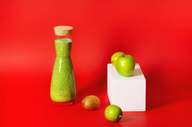 Bir şişe yeşil karışık meyve suyu kırmızı zemin üzerinde duruyor, yanında yeşil meyveli beyaz geometrik prizma uzanıyor. Yatay yönelim.