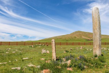 Standing deer stones in on a Mongolian hill, with a red fence in the bacck ground and blue skies clipart