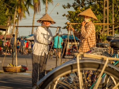 Konik şapkalı iki Vietnamlı kadın, öğleden sonra nehrin kıyısında sırık taşıyorlar, Hoi An, Vietnam - 10 Ocak 2015