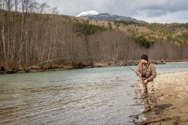 Kalum Nehri, British Columbia, Kanada 'da büyük bir balığa kancayı takmış bir balıkçı. Uzak kıyılarda kamış ve kavak ağacı ve arka planda dağlar var.