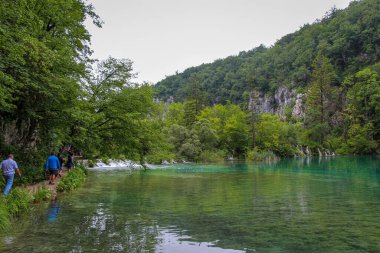 Plitvice Gölleri, Hırvatistan - 14 Temmuz 2018: Hırvatistan 'ın UNESCO Dünya Mirasları Bölgesi' nde, arka planda şelaleler bulunan sığ bir göl kenarında yürüyen turistler