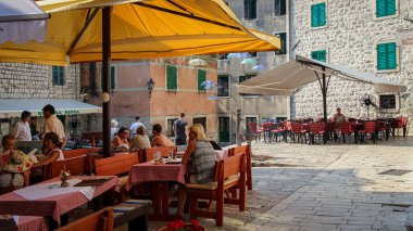 Eski Kotor kentinde bir meydanda, arka planda uçan şemsiyeler bulunan bir kafe, Karadağ