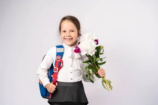 first grade school girl in a white shirt holding flowers and a backpack