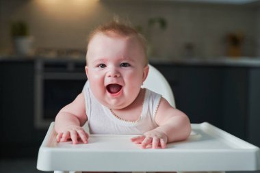 Beyaz elbiseli küçük bir çocuk yüksek bir sandalyede oturur ve gülümser..