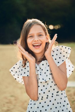 Plajda puantiyeli bir elbise giymiş küçük bir kız gülüyor ve ellerini yana açıyor.