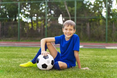 Küçük bir çocuk yeşil bir futbol sahasında oturuyor, bir futbol topu..