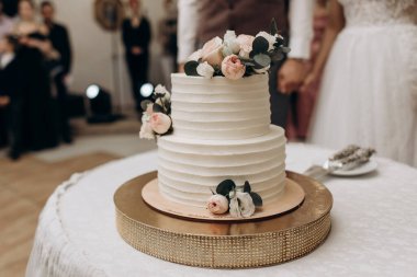 Beyaz masanın üzerinde yeşil yapraklar, pembe ve beyaz güllerle süslenmiş beyaz iki katlı düğün pastası.