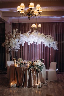 Restoran salonundaki şenlik masasında beyaz ve pembe gül buketleri, beyaz zambaklar, yanan mumlar ve masanın üstünde beyaz bir çiçek kemeri. Lokantada tatil masası servis edilmiş. Beyaz masa örtüleriyle kaplı ziyafet masası.