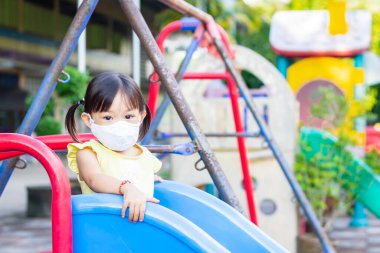 Mutlu Asyalı kız çocuğu gülümsüyor ve kumaş maske takıyor. Çocuk parkında, sosyal mesafede sürgülü bar oyuncağıyla oynuyor. Çocuk konseptini öğrenmek ve aktif olmak.