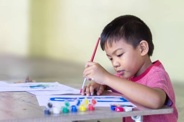 Seçici odaklanma. 5 yaşındaki bir çocuğun portresi. Asyalı öğrenci odadaki kağıda renkler çiziyor ve boyuyor. Evden öğrenim, sosyal uzaklık, çocuk ve eğitim kavramı.