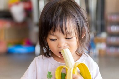2 yaşındaki bir bebeğin portresi. Mutlu Asyalı kız çocuğu, tatlı bir gülümsemeyle muz yiyip ısırmaktan zevk alıyor. Yiyecek ve sağlıklı çocuk konsepti.