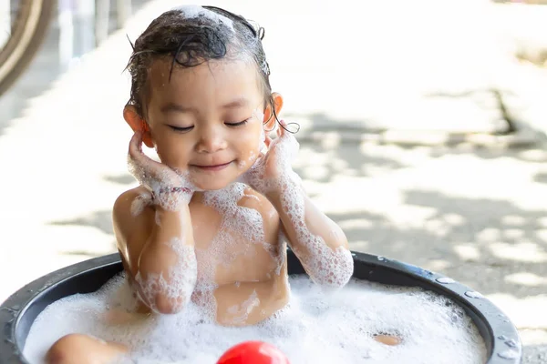 2 yaşındaki bir bebeğin portresi. Mutlu Asyalı bebek köpüklü sabunla banyo yapıyor. Küvette oturuyor ve bir top oyuncağıyla oynuyor. Tatlı gülümseme. Beyaz arkaplanda izole.
