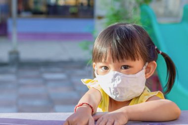 2-3 yaşlarındaki bir bebeğin portresi. Mutlu Asyalı kız çocuğu gülümsüyor ve kumaş maske takıyor. Çocuk parkını işaret ediyor. Sosyal uzaklık. Çocuk konseptini öğrenmek ve aktif olmak.