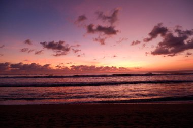 Kuta plajında dramatik bir gün batımı manzarası. Mor, turuncu ve mavi gökyüzü.