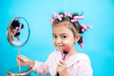 Saçında bigudilerle gülümseyen küçük bir kız mavi izole bir arkaplanda kameraya bakıyor. Bigudili küçük bir kız el aynasında tarak ve allık için fırça tutuyor. Metin için boş boşluk.
