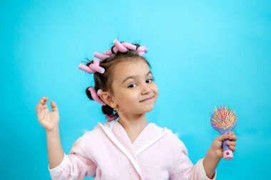 Saçında bigudilerle gülümseyen küçük bir kız mavi izole bir arkaplanda kameraya bakıyor. Bigudili küçük bir kız el aynasında tarak ve allık için fırça tutuyor. Metin için boş boşluk.