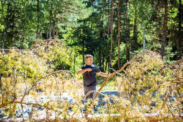 A young European man takes a photo with a selfie stick while standing on a fallen tree over a mountain river around a pine forest. A young man holds a selfie stick with a camera in front of him in a pine forest.