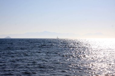 Açık Ege Denizi tam güneş altında, bir tekne uzaklarda yol alıyor. Arkaplanda dağlar