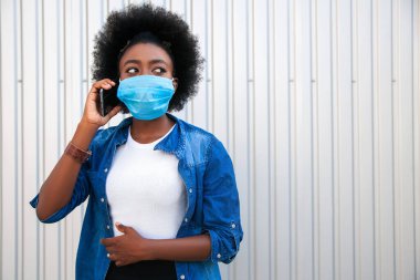 Sağlık sigortası konsepti. Gündelik giyinmiş, tek kullanımlık tıbbi maske takan, gri arka planda akıllı telefon kullanan genç bir Afrikalı kadın. Boşluğu kopyala.