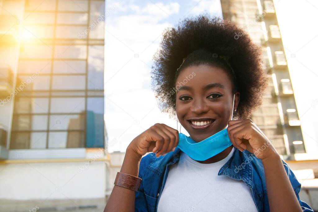 Quarantine is over, coronavirus is finish. Young beautiful black african american woman with curly black hair taking her medical protective mask off. City background.