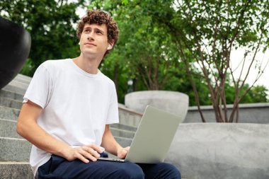 Eğitim, teknoloji ve internet kavramı - Avrupa 'lı genç erkek öğrenci kablosuz kulaklıklar ve dizüstü bilgisayarla yaz günü şehir parkında eğitim görüyor ve müzik dinliyor. Boş boşluk.