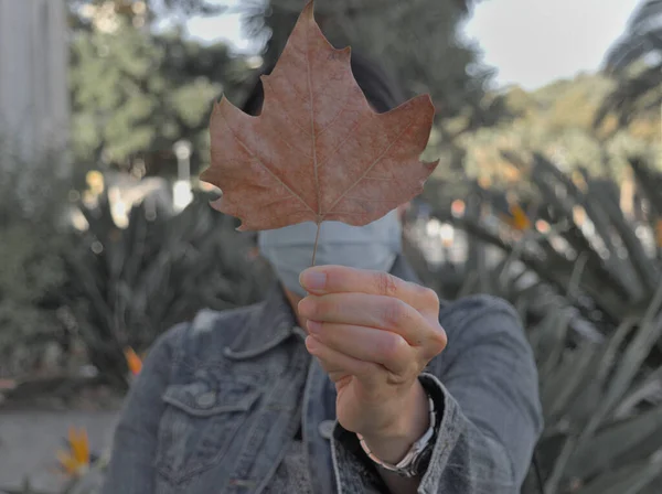 A person with a medical mask holding an autumn leave in a green background