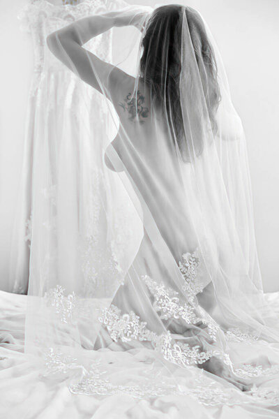 Naked bride covered in veil poses with her back to the camera in the background of the wedding dress.