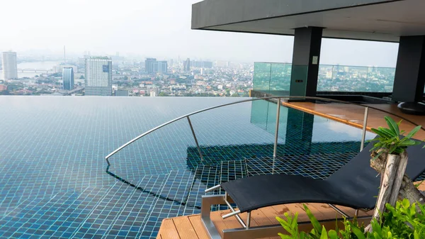There is ledge lounger in the corner of the pool and the background there are lots of building in the city with air pollution.