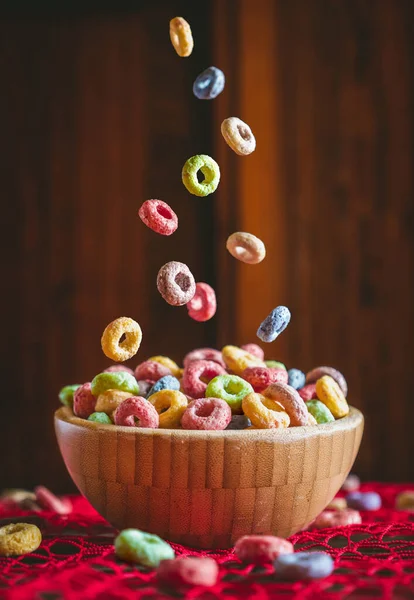 Colorful round fruit cereals falling into a wooden bowl on a red table. Preparing breakfast.