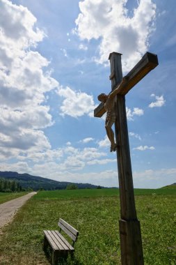 Kahverengi ahşap çarmıha gerilmiş İsa figürü. Tahtadan yapılmış bir koltuk, daha dar bir patika yeşil çayırlardan geçiyor. Beyaz bulutlu mavi gökyüzü. Almanya.