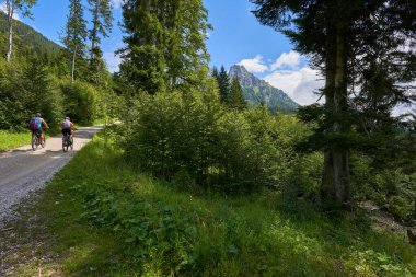 Tannheimer dağ papağanı başı, parlak çakıl taşlı patika yüksek bir karaya doğru gidiyor, iki bisikletçi yolda, mavi gökyüzü beyaz bulutlu. Bavyera, Yüzeysel.