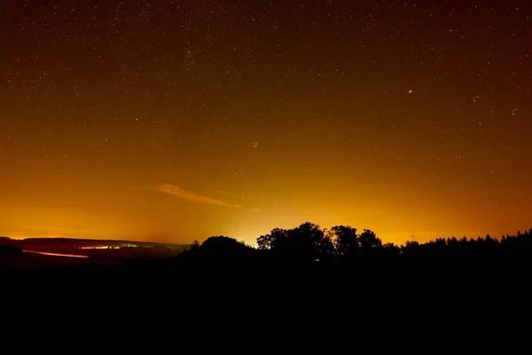 Turuncu gece gökyüzü birçok takım yıldızıyla, Mars gezegeni görülebilir, ön plandaki orman bir siluet olarak gösterilir. Uzun pozlama hareketli bir arabayı kırmızı çizgi olarak gösteriyor. Almanya. Heroldstatt.