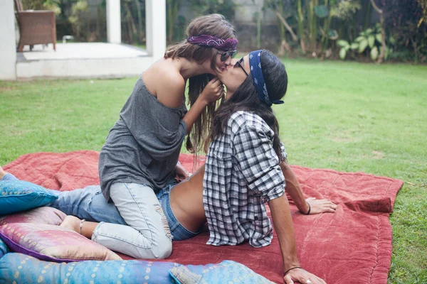Attraktiver junger Mann, der auf einer Decke im Gras sitzt. schöne Mädchen sitzt oben auf Kerl und küsst ihn. Hippie- und Zigeunerstil. — Stockfoto