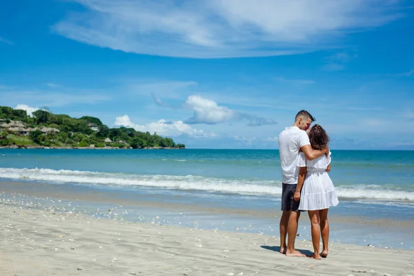 Vista trasera de pareja romántica joven en ropa blanca de pie en la playa tropical. Hombre y mujer abrazándose en el lado izquierdo de la foto. Cielo azul y ver en el fondo — Foto de Stock
