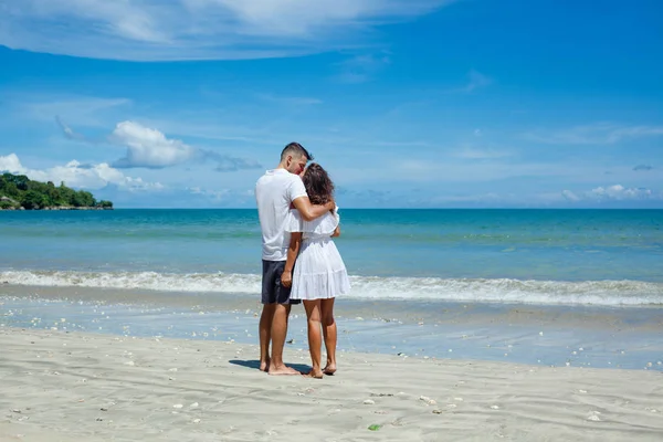 Vista trasera de pareja romántica joven en ropa blanca de pie en la playa tropical. Hombre y mujer abrazándose en medio de la foto. Cielo azul y ver en el fondo — Foto de Stock