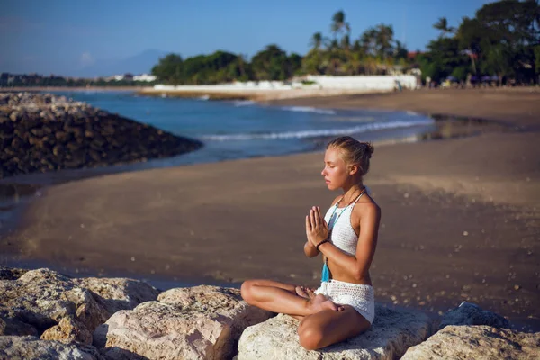 Νεαρή όμορφη γυναίκα σε λευκό μπικίνι πρακτική γιόγκα κάθεται στην ακτή της θάλασσας πέτρα με τροπική θέα στο παρασκήνιο. Κάθεται στη θέση του λωτού. Οι παλάμες της είναι διπλωμένες για προσευχή. Διαλογισμός — Φωτογραφία Αρχείου