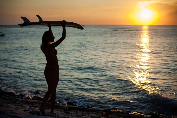 Silueta dívky v podsvětlici, která držela SURFOVOU desku nad hlavou. Západ slunce na mořském pobřeží — Stock fotografie
