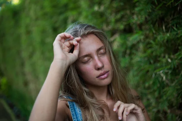 Nära porträtt av en ung kvinna utan smink med slutna ögon. En flicka står mot en grön gräs vägg och håller händerna bredvid huvudet. Ömhet och naturlighet — Stockfoto