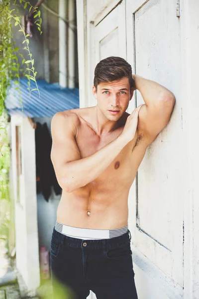 Portrait of a beautiful sexy brunette with nude sport torso in black jeans. a young man stands outside leaning on a white wall. relief biceps and press