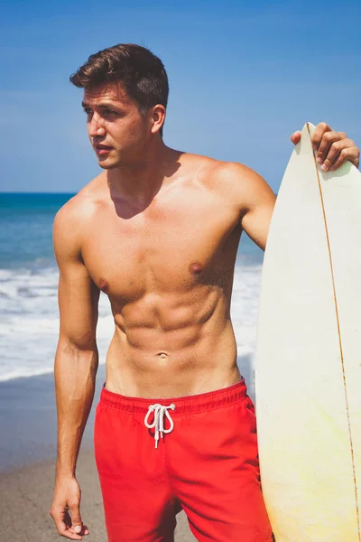 Kırmızı şortlu seksi çekici bronzlaşmış genç adam ve plajda bir sörf tahtası ile çıplak göğüslü uygun