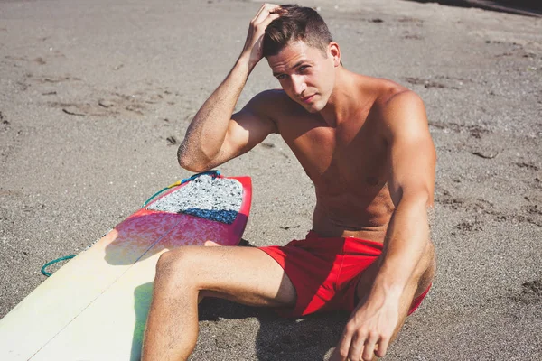 Nahaufnahme Porträt eines sexy, sehr attraktiven, braun gebrannten jungen Mannes in roten Shorts und nacktem Oberkörper, der am Strand sitzt. Surfer, Modell, Kerl, Sand — Stockfoto