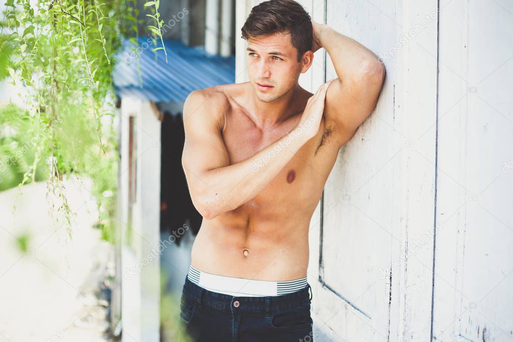 Portrait of a beautiful sexy brunette with nude sport torso in black jeans. a young man stands outside leaning on a white wall. relief biceps and press. sunny day