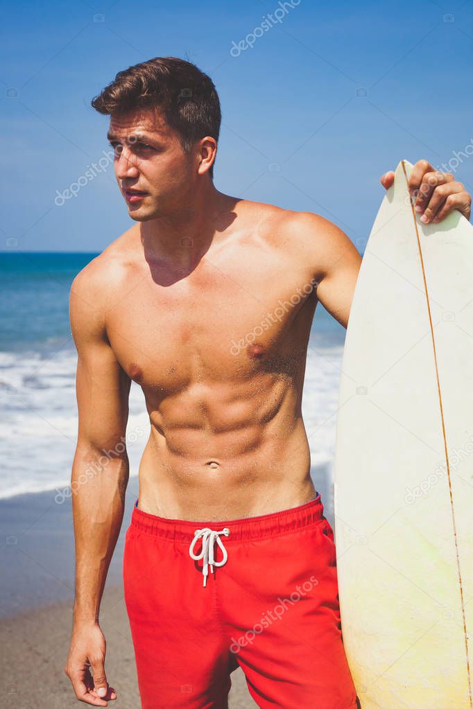 Sexy attractive tanned young man in red shorts and fit bare-chested with a surfboard on the beach