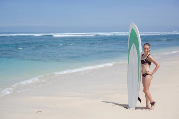 Na pláži stojí krásná usmívající se mladá žena v bikinách se surfovým prkněm. slunné denní modré moře — Stock fotografie