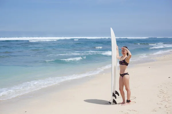 Krásná usměvavá mladá žena v bikinách se surfovým prkněm stojí na pláži a dívá se do dálky. slunečný den modrý oceán — Stock fotografie