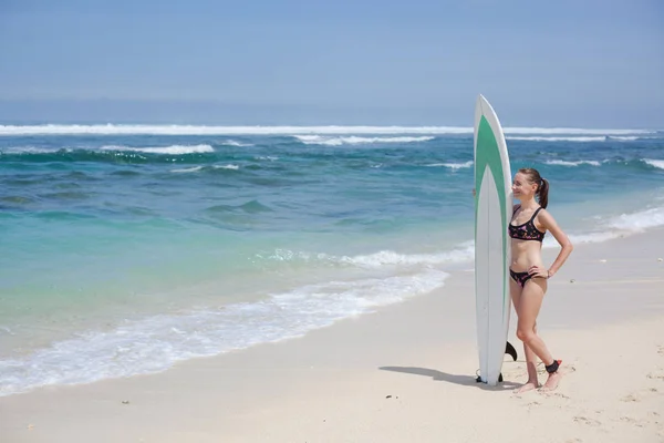 Krásná usměvavá dívka s příbojem v bikinách se surftem stojí na pláži a dívá se do dálky. slunečný den modrý oceán a obloha — Stock fotografie