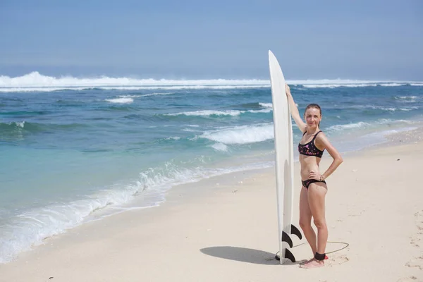 Krásná sexy usměvavá mladá žena v bikinách se surfovým prkněm stojí na pláži. slunné denní modré moře — Stock fotografie