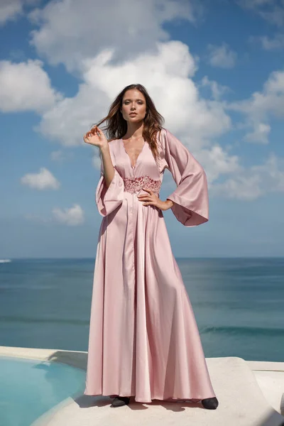 Belle jeune femme modèle dans une longue robe rose soie se dresse contre le ciel bleu et vue sur la mer. pose de mode. nuages blancs en arrière-plan — Photo